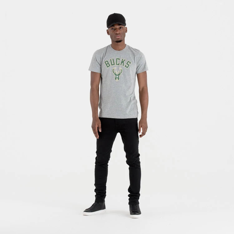 Milwaukee Bucks NBA Regular Grey T-Shirt, on-model view showing a grey T-shirt with "BUCKS" lettering and deer logo in green on the front, worn by a person standing with a black cap and black jeans.