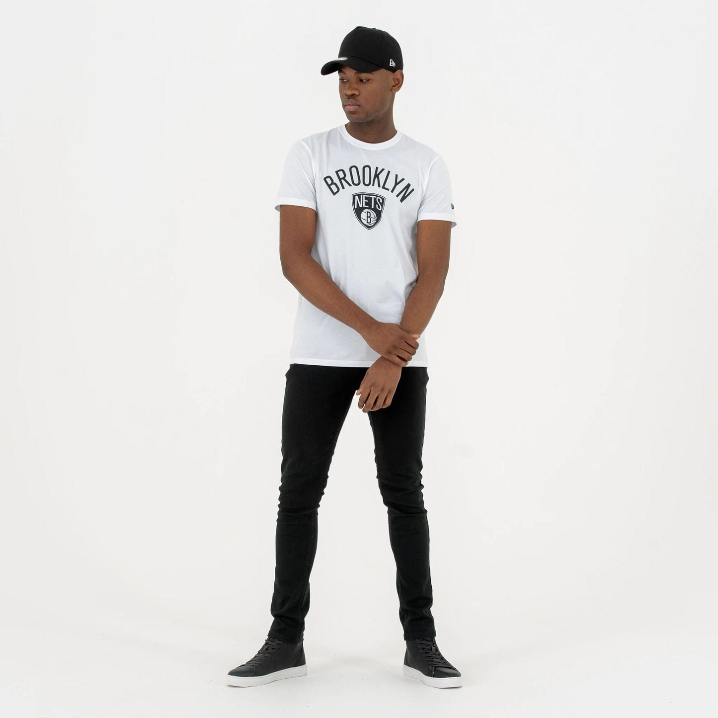 Brooklyn Nets NBA Regular White T-Shirt, on-model view showing the black Brooklyn Nets logo centred on the chest, with a small raised New Era logo on the side of a black cap worn by the model.