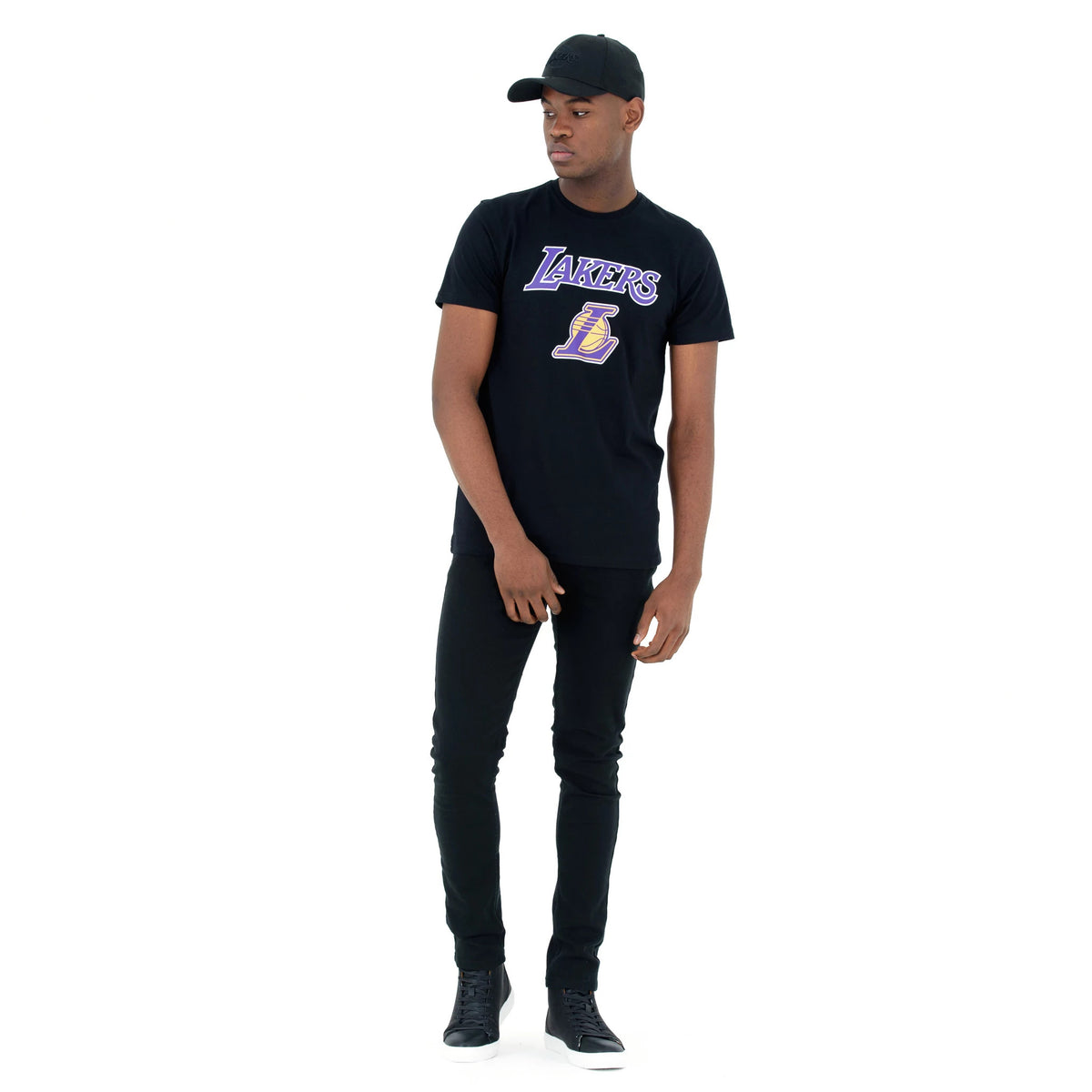 LA Lakers NBA Regular Black T-Shirt, on-model view showing a black T-shirt with a large purple and gold Lakers logo across the chest, paired with black trousers and a black cap featuring a raised New Era logo.