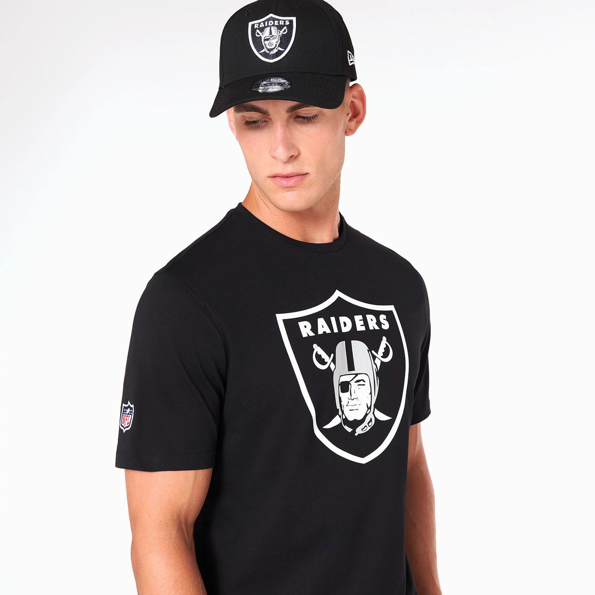 Las Vegas Raiders NFL Black T-Shirt, on-model view showing the large white Raiders logo printed on the chest and a small embroidered NFL logo on the left sleeve, paired with a black cap featuring a raised Raiders logo on the front.