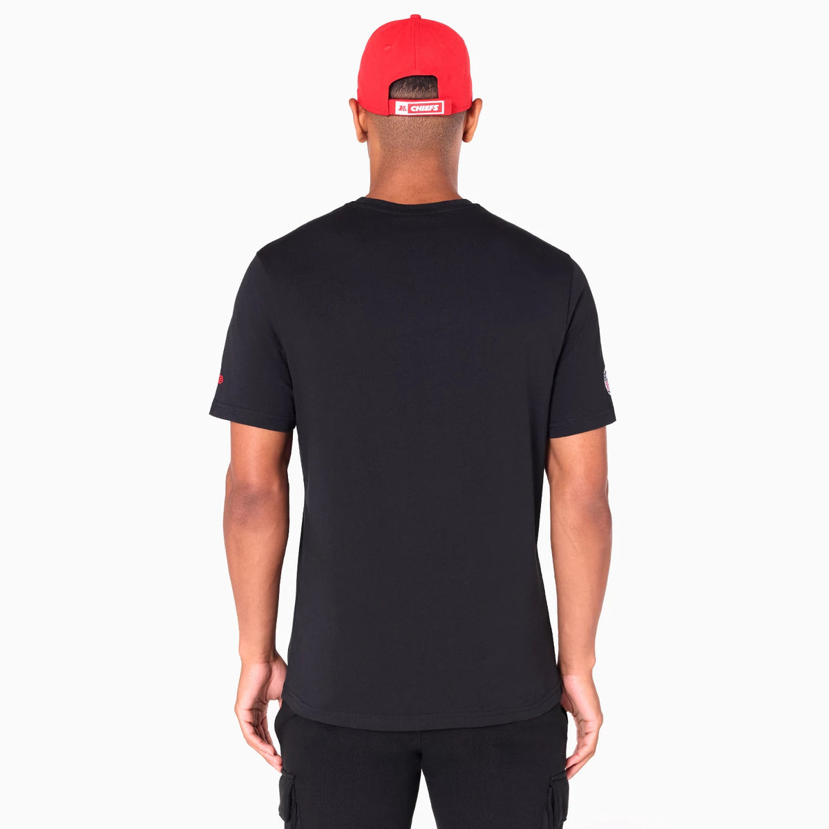 New Era Kansas City Chiefs NFL Black T-Shirt, rear view showing the plain back of the black T-shirt worn by a person, with a red cap displaying a logo patch above the adjustable strap at the back.