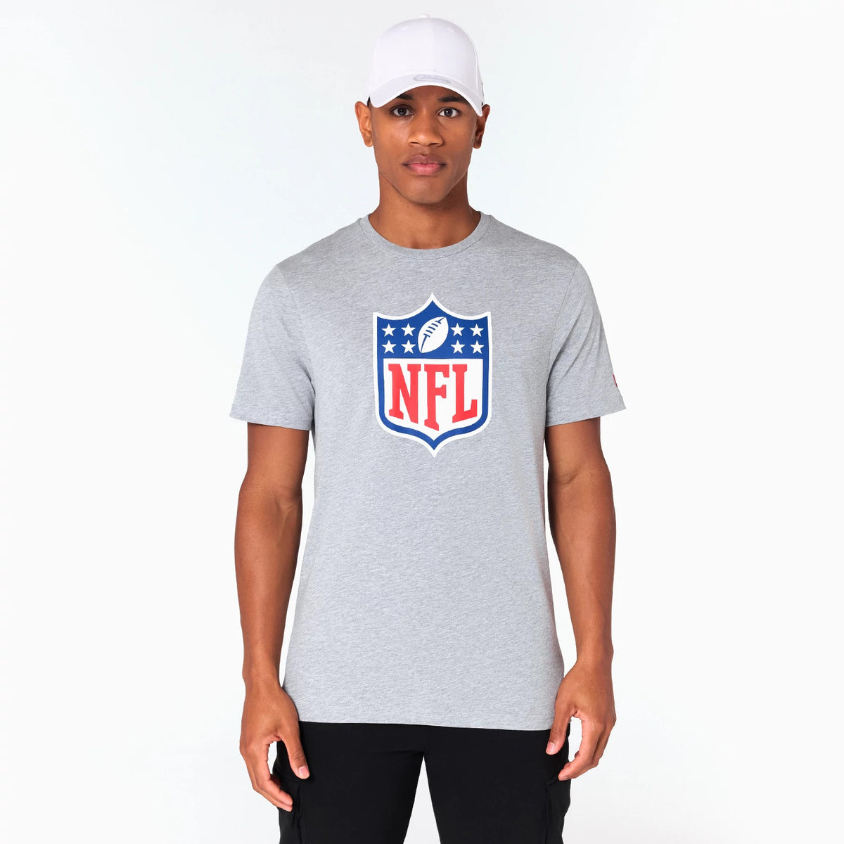 New Era NFL Shield Logo NFL Grey T-Shirt, on-model view showing the grey tee with a large shield design in red, white, and blue on the chest, featuring the NFL lettering prominently, worn by a person with a white cap and black trousers.