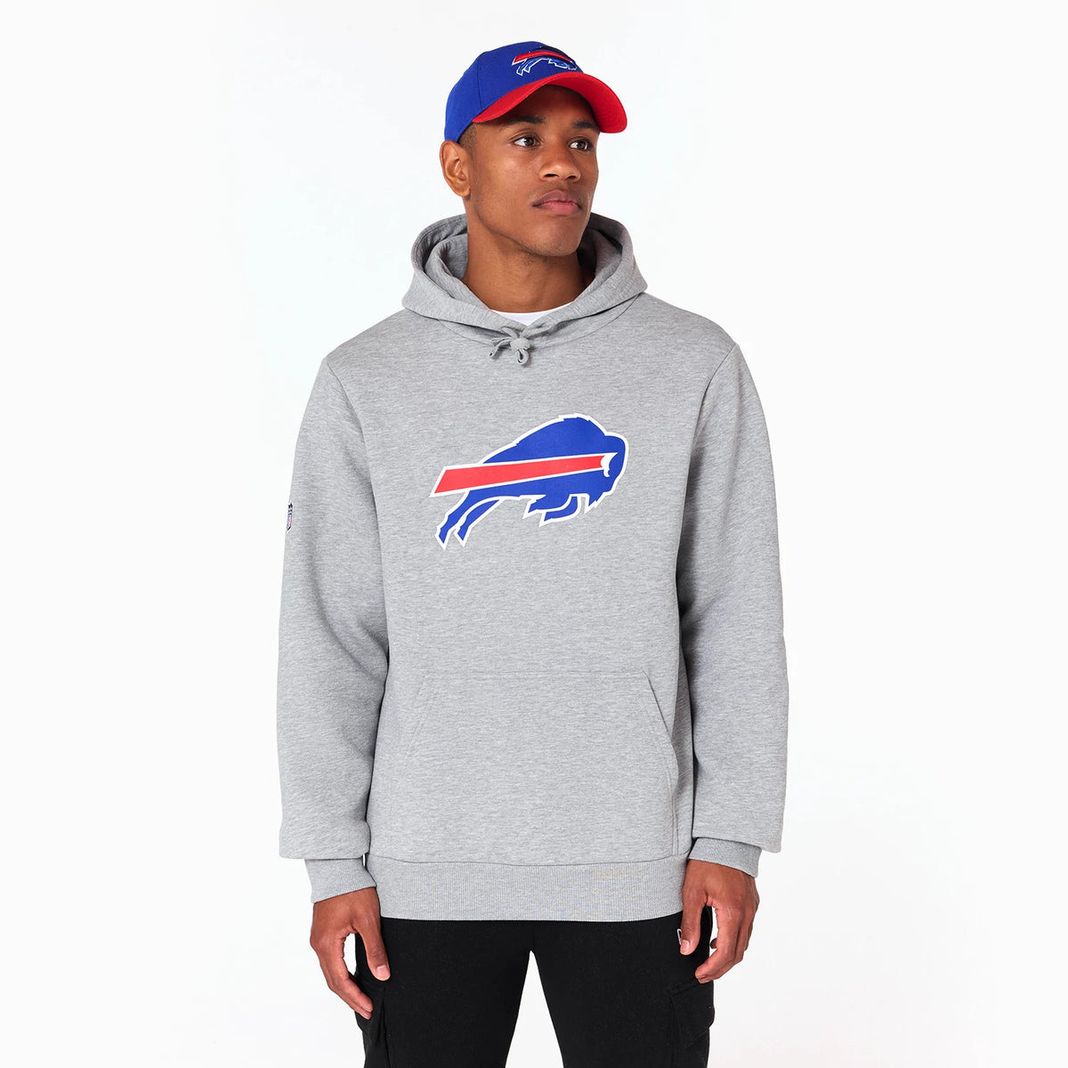 New Era Buffalo Bills NFL Grey Pullover Hoodie, on-model view showing the grey hoodie with a large buffalo emblem on the chest, a drawstring hood, and a debossed NFL logo on the left sleeve, worn with a blue and red cap.
