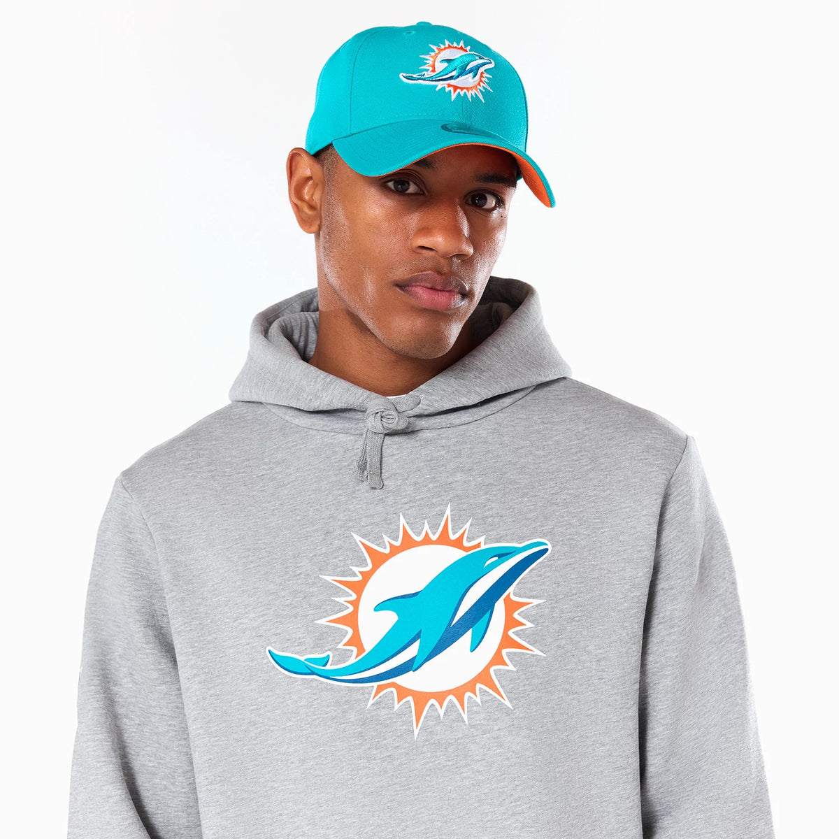 New Era Miami Dolphins NFL Grey Pullover Hoodie, on-model view featuring a large turquoise and orange logo printed on the front, a drawstring hood resting on the shoulders, and the model wearing a turquoise cap with a raised logo on the front panel.
