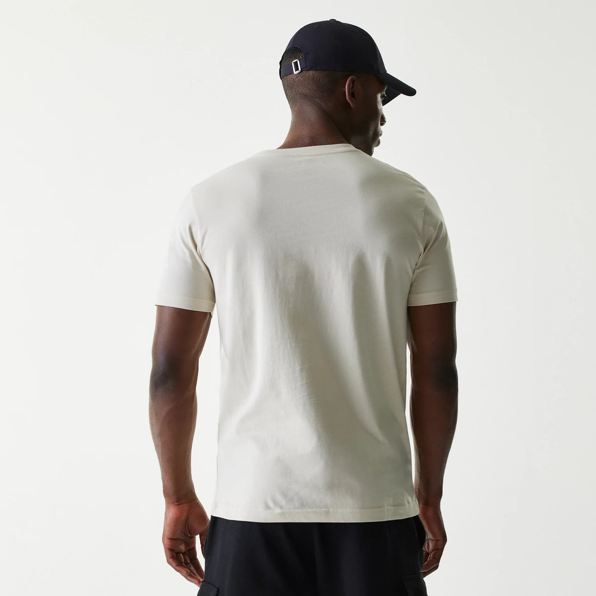 New Era LA Dodgers MLB City Connect Cream T-Shirt, rear view of a cream T-shirt worn by a person, showing a simple back design with short sleeves and a plain neckline, paired with a dark cap featuring a metal buckle on a fabric strap.