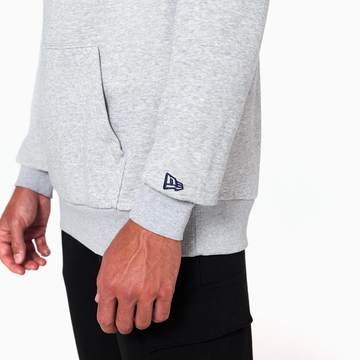 New Era Washington Wizards NBA Grey Pullover Hoodie, side view showing the light grey fabric with a front pocket and a raised dark blue New Era logo on the left sleeve cuff, part of a casual outfit with black trousers.