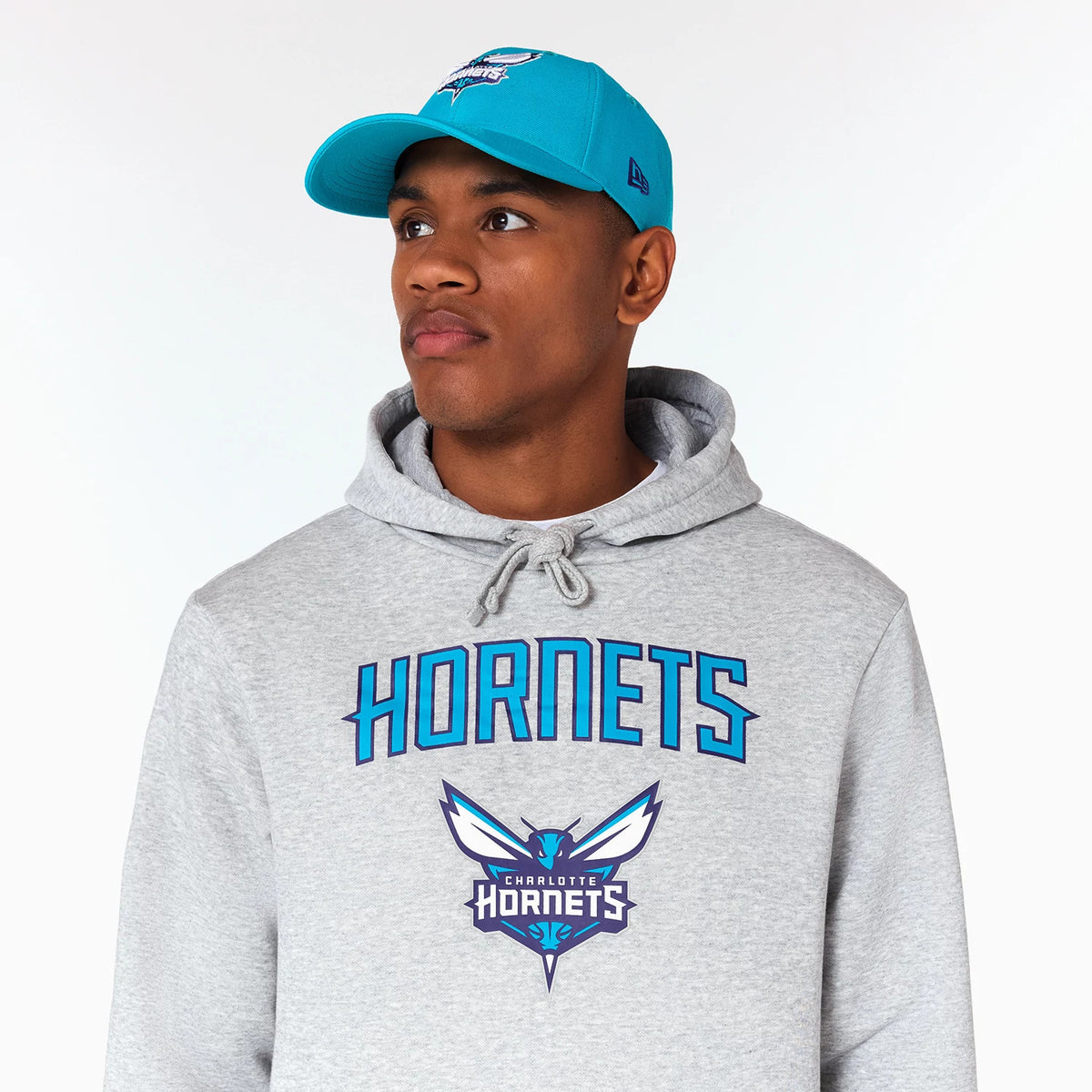 New Era Charlotte Hornets NBA Grey Pullover Hoodie, on-model view showing the grey hoodie with a large embossed team logo on the chest, blue HORNETS text above and a matching blue cap worn by the model.
