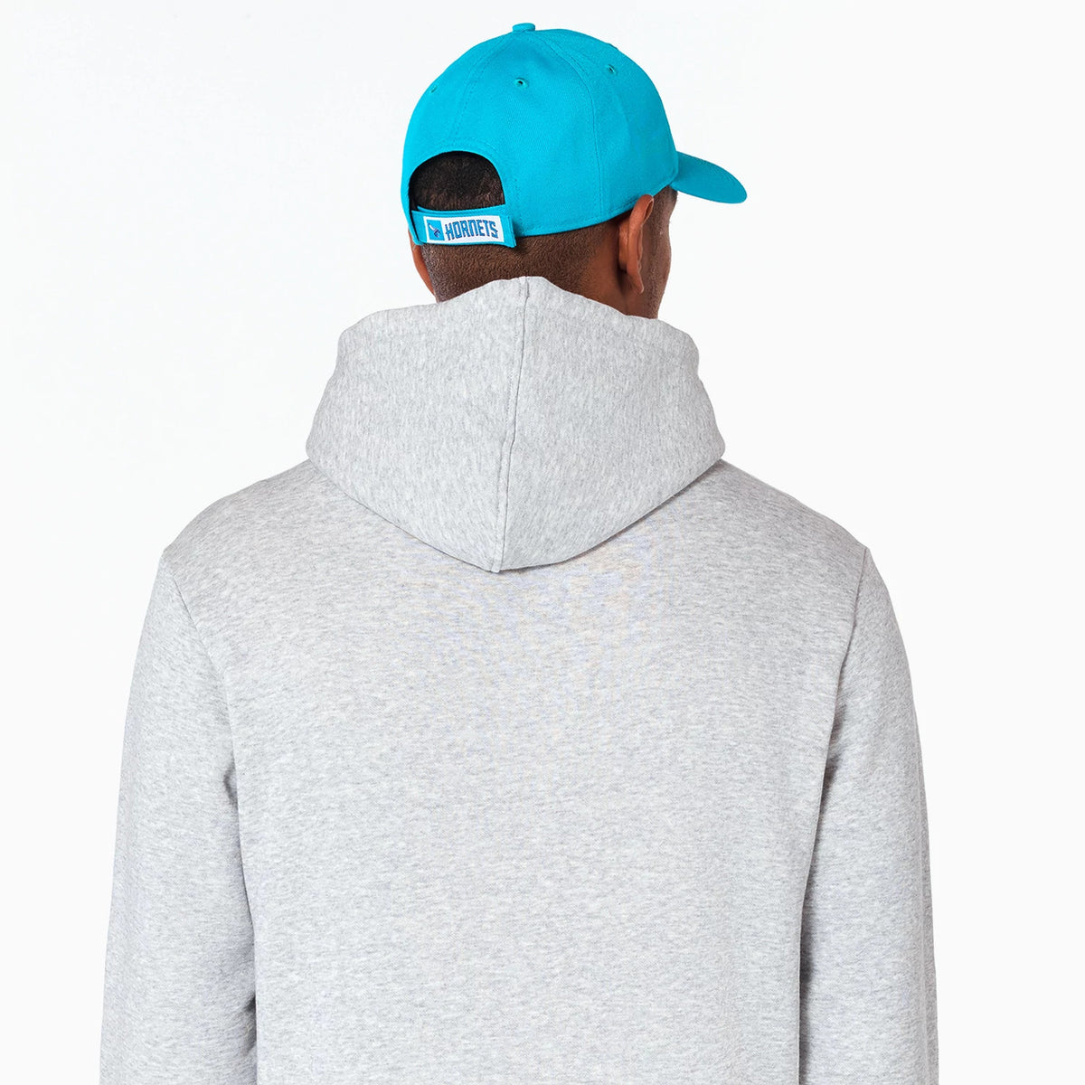 New Era Charlotte Hornets NBA Grey Pullover Hoodie, rear view showing the light grey hood against the back, worn by a person in a blue cap with an embossed Hornets logo on the adjustable strap.