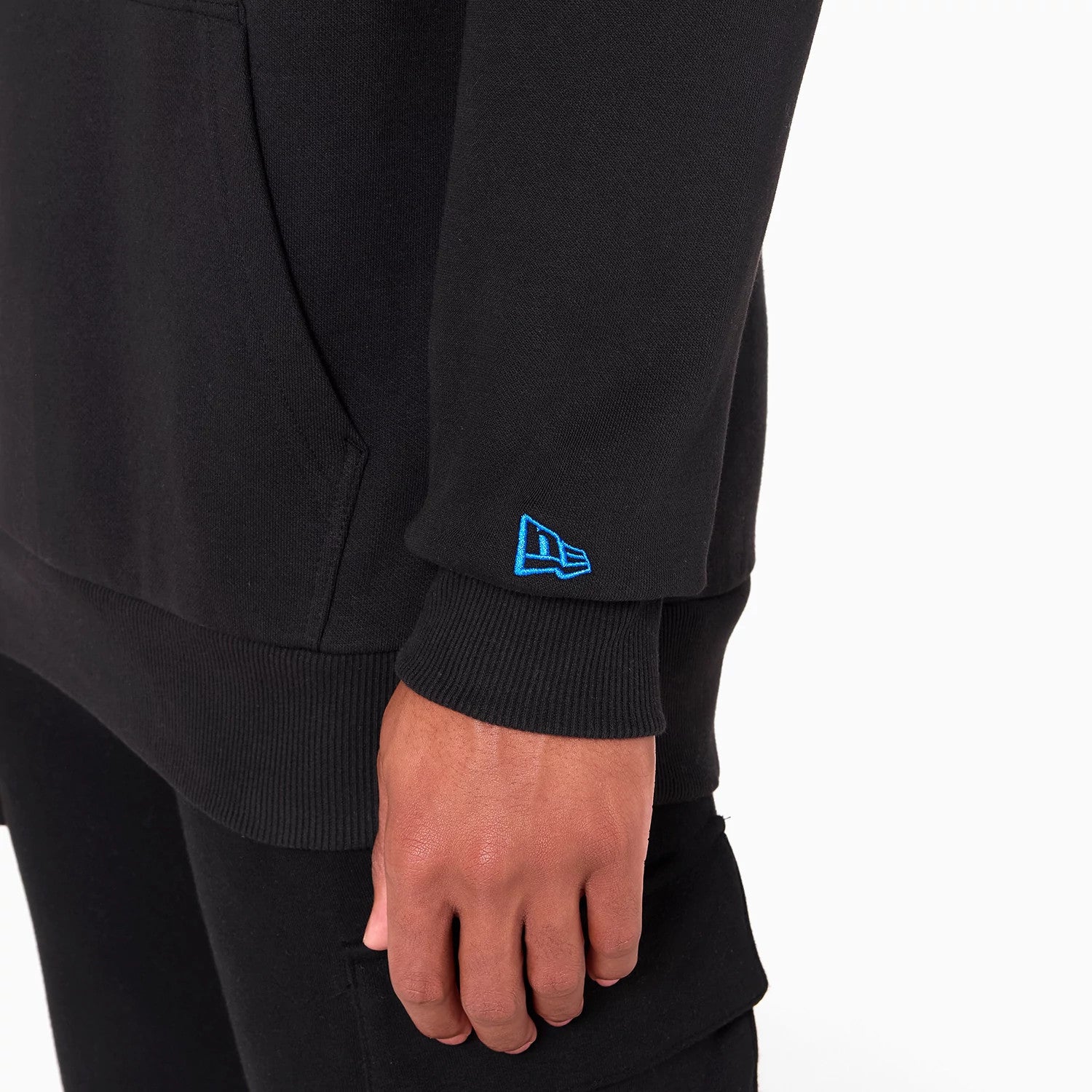 New Era Orlando Magic NBA Black Pullover Hoodie, side view showing the lower half with a blue embroidered New Era logo on the cuff, ribbed hem at the wrist, and the edge of a kangaroo pocket on the black fabric.