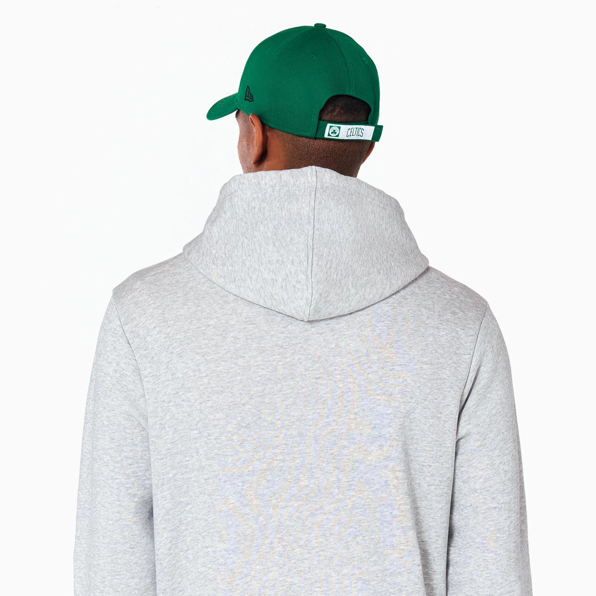 New Era Boston Celtics NBA Grey Pullover Hoodie, rear view showing the grey fabric and hood from behind, paired with a green cap featuring a debossed New Era logo on the side and an embroidered Celtics logo on the back strap.