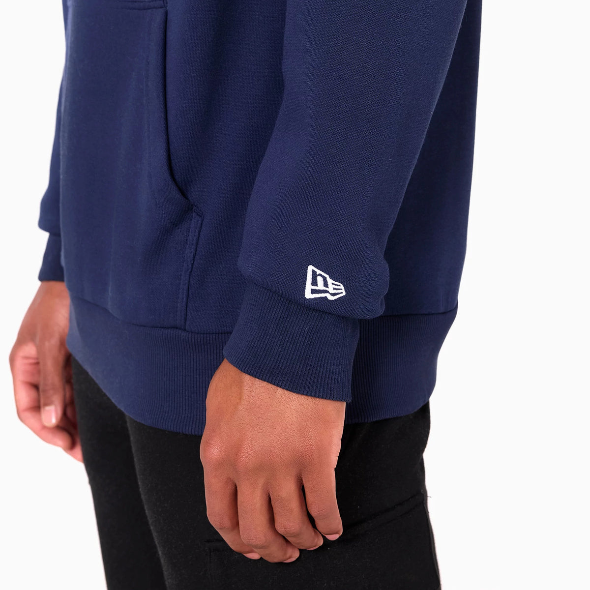 New Era Utah Jazz NBA Navy Pullover Hoodie, side view showing the lower half with a white embroidered New Era logo on the sleeve cuff, a spacious front pocket, and a ribbed hem above black trousers worn by a model.