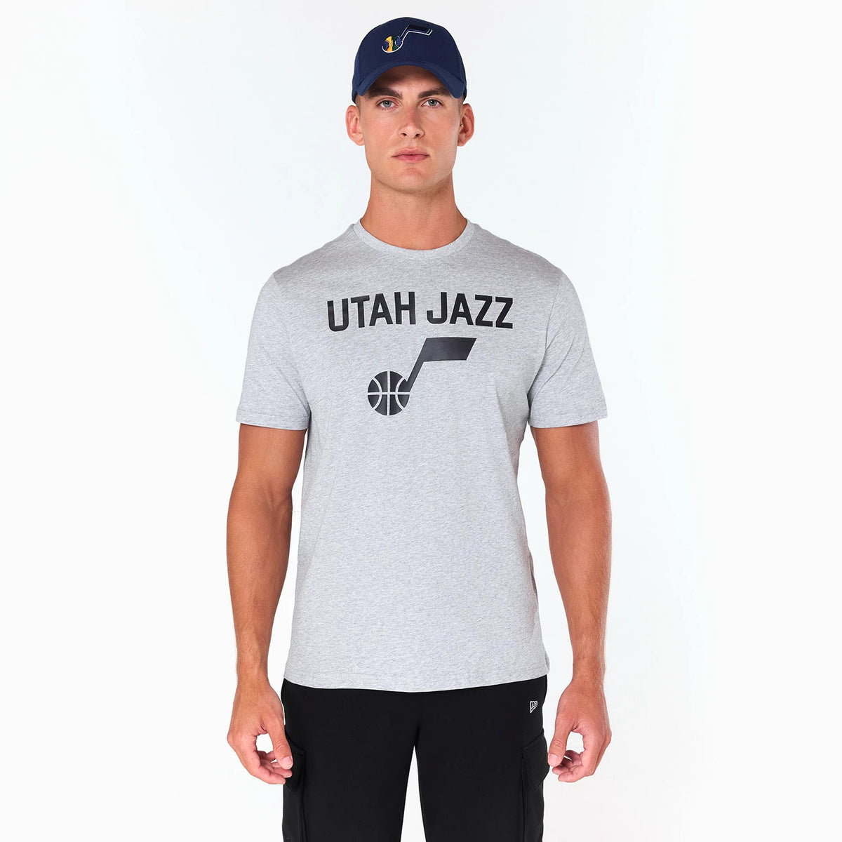 New Era Utah Jazz NBA Grey T-Shirt, on-model view showing the grey T-shirt with black 'Utah Jazz' text and logo printed on the chest, complemented by a raised logo on a dark blue cap worn by the model.