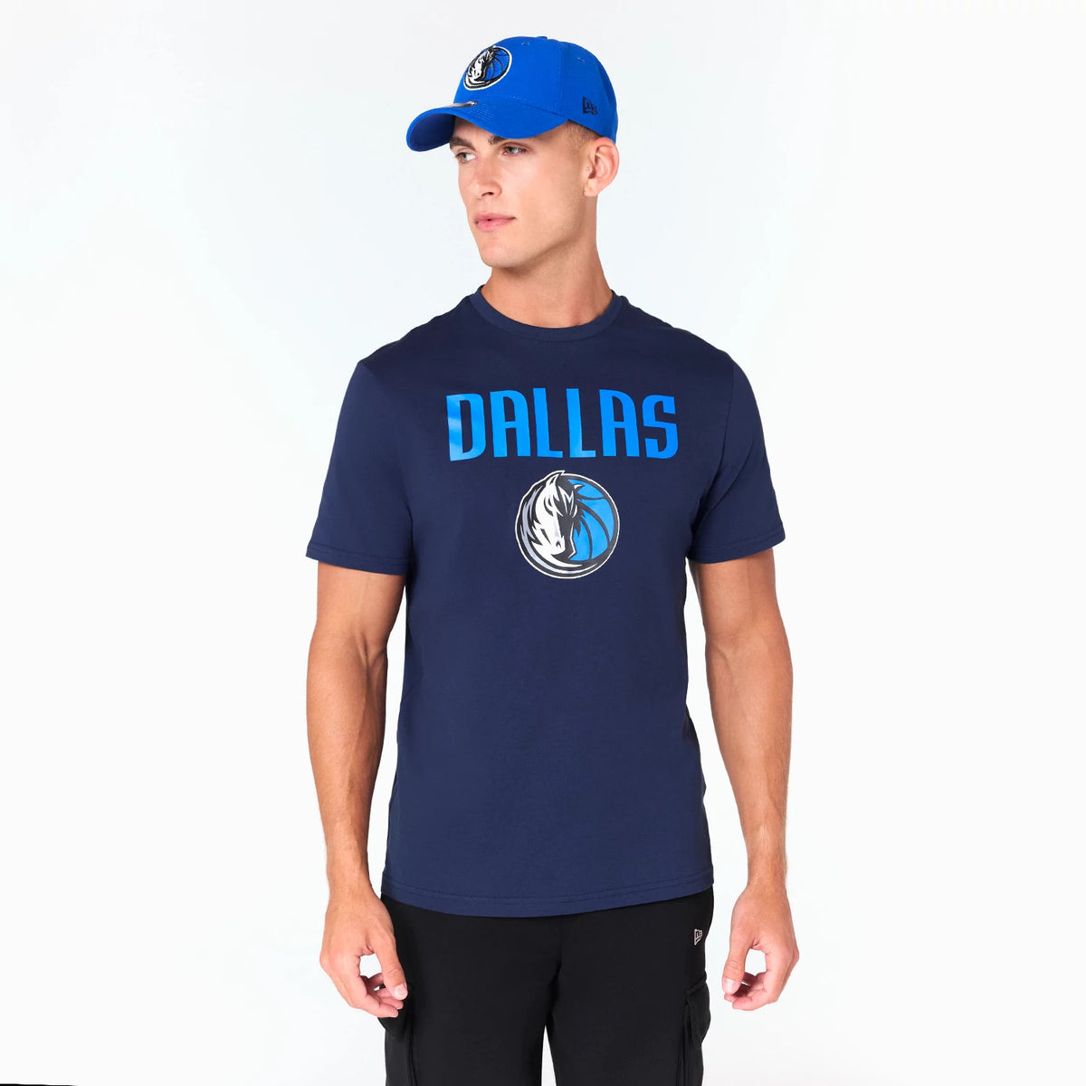 New Era Dallas Mavericks NBA Navy T-Shirt, on-model view showing a navy shirt with DALLAS in blue letters and the Mavericks logo on the chest, paired with a blue cap featuring an embroidered Mavericks logo on the front.
