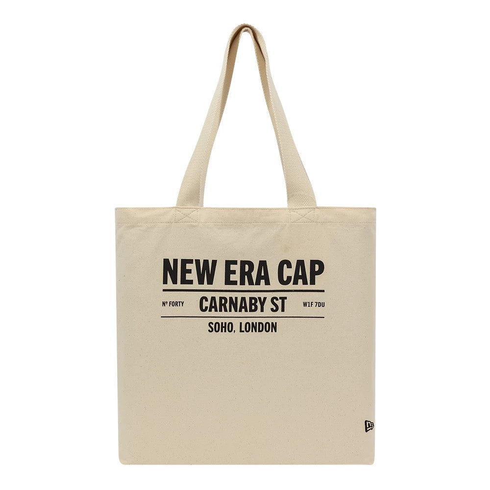 New Era Cap Carnaby Address Light Beige Canvas Tote Bag, front view showing bold black text displaying New Era Cap and Carnaby St address, with dual shoulder straps extending from the top and a small debossed New Era logo in the bottom right corner.