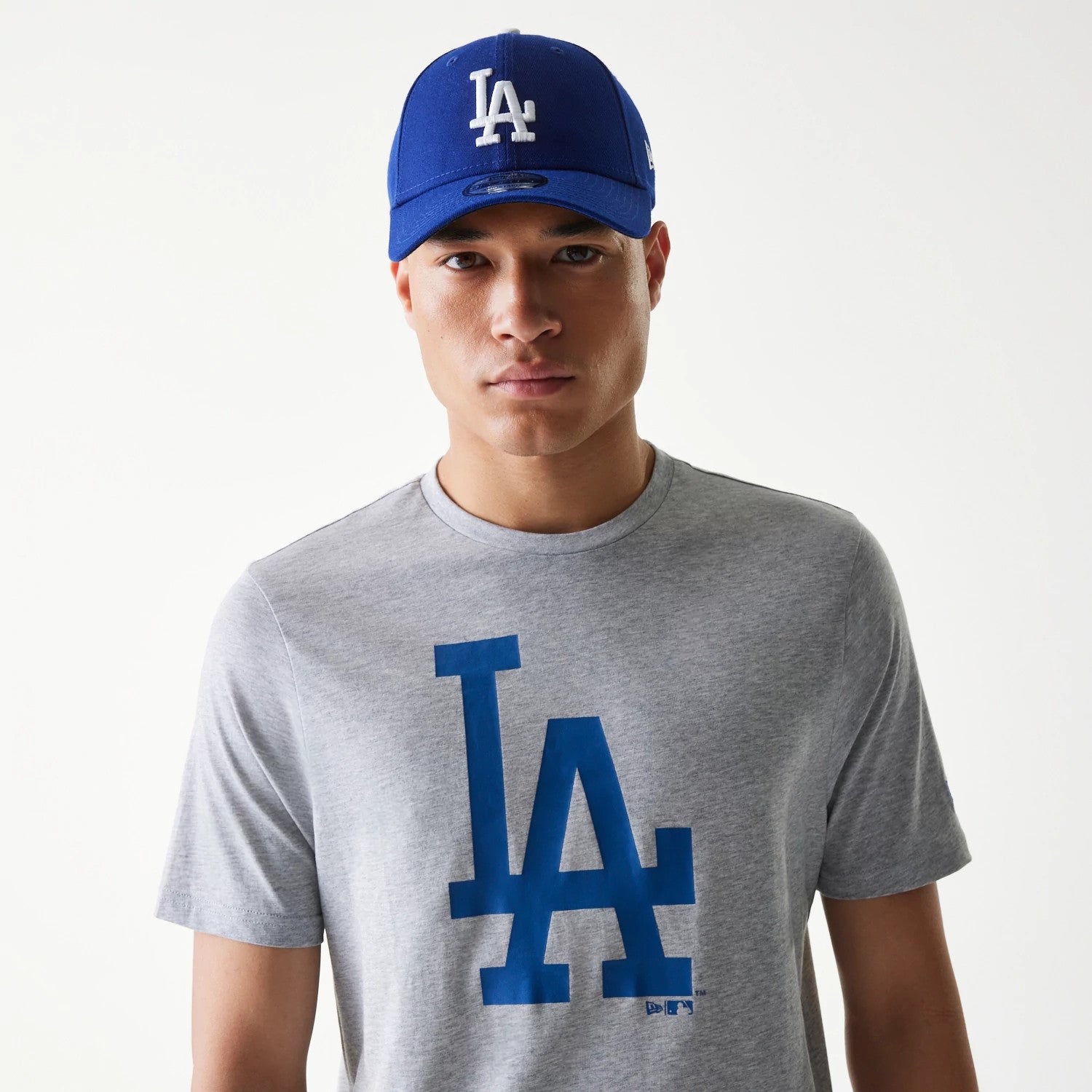 New Era LA Dodgers MLB Regular Grey T-Shirt, on-model view featuring a large blue LA logo centred on the chest, complemented by a blue cap with a raised white LA logo, eyelets on top, and a grey background.