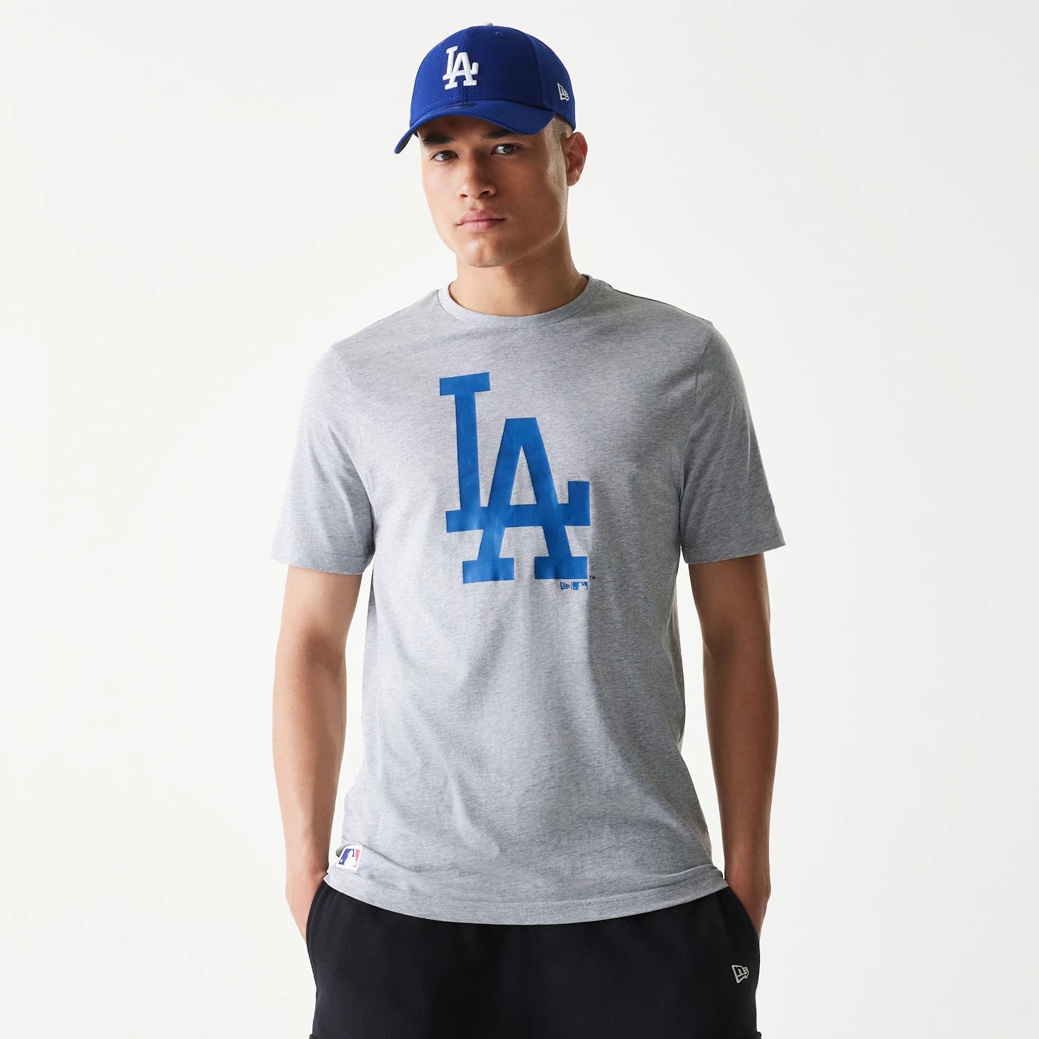 New Era LA Dodgers MLB Regular Grey T-Shirt, front view showing a large blue LA logo on the chest, worn by a person also wearing a blue cap with a white LA logo on the front and a debossed New Era logo on the side of the cap.