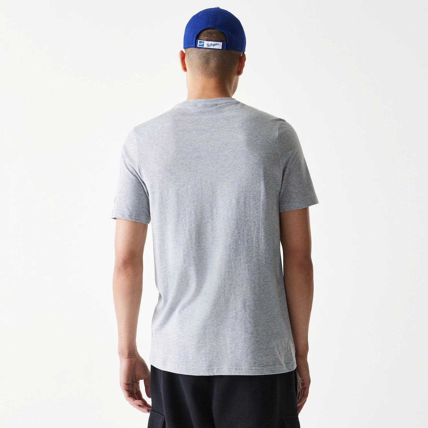 New Era LA Dodgers MLB Regular Grey T-Shirt, rear view showing the plain grey back of the T-shirt worn by a person, paired with dark shorts and a blue cap featuring a debossed rectangular logo at the back.