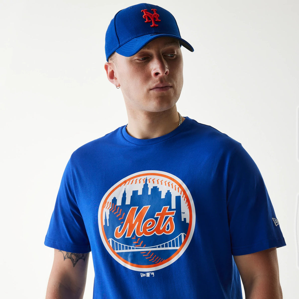 New Era New York Mets MLB Regular Blue T-Shirt, on-model view showing a large orange and white Mets print on the front, the sleeve featuring an embroidered New Era logo, worn with a matching blue cap with a raised orange Mets logo.