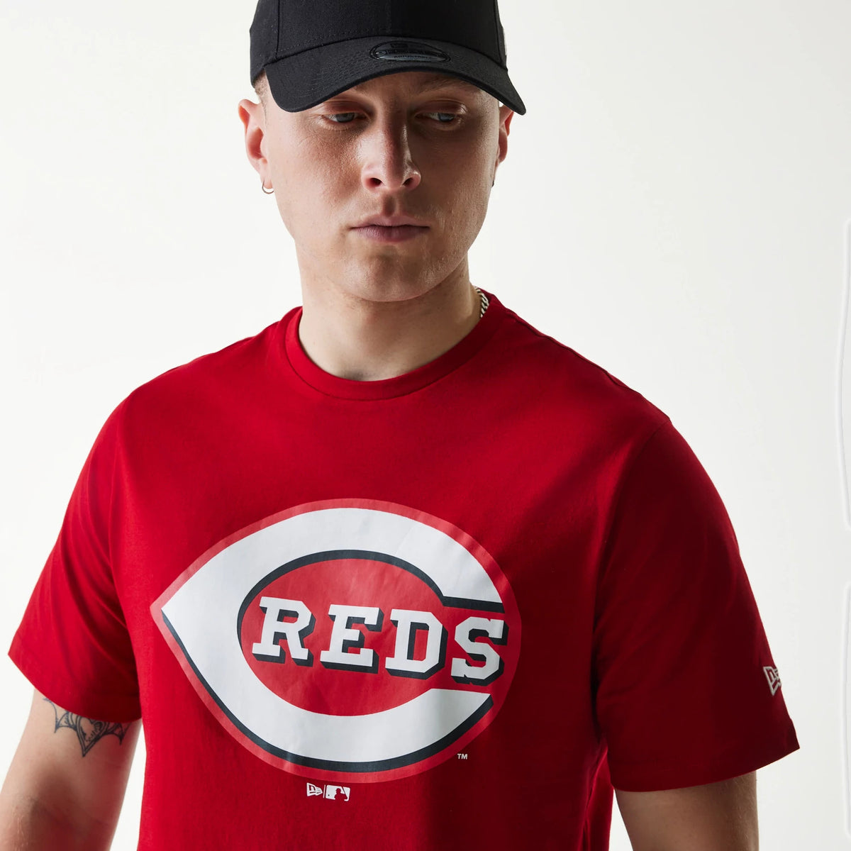 New Era Cincinnati Reds MLB Regular Red T-Shirt, on-model view showing a large Cincinnati Reds logo printed in white and black on the chest, with a raised New Era logo on a black cap worn by the model looking to the side.