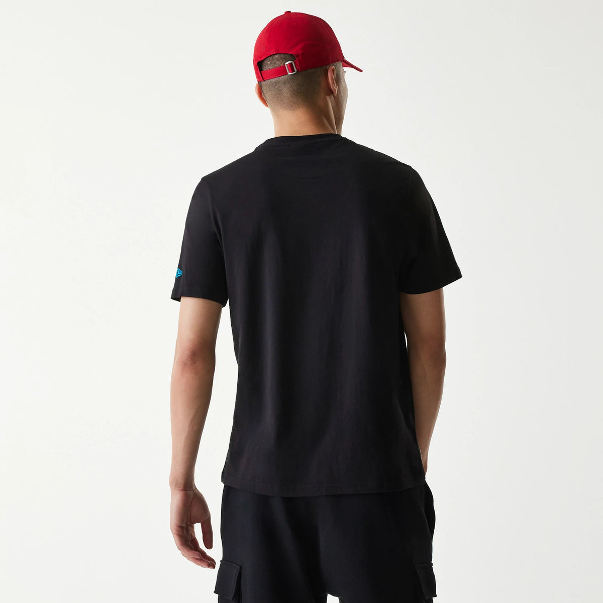 New Era Miami Marlins MLB Regular Black T-Shirt, rear view showing the plain black back with short sleeves, a blue embossed logo on the left sleeve, worn by a person in black shorts and a red cap with a buckle strap.