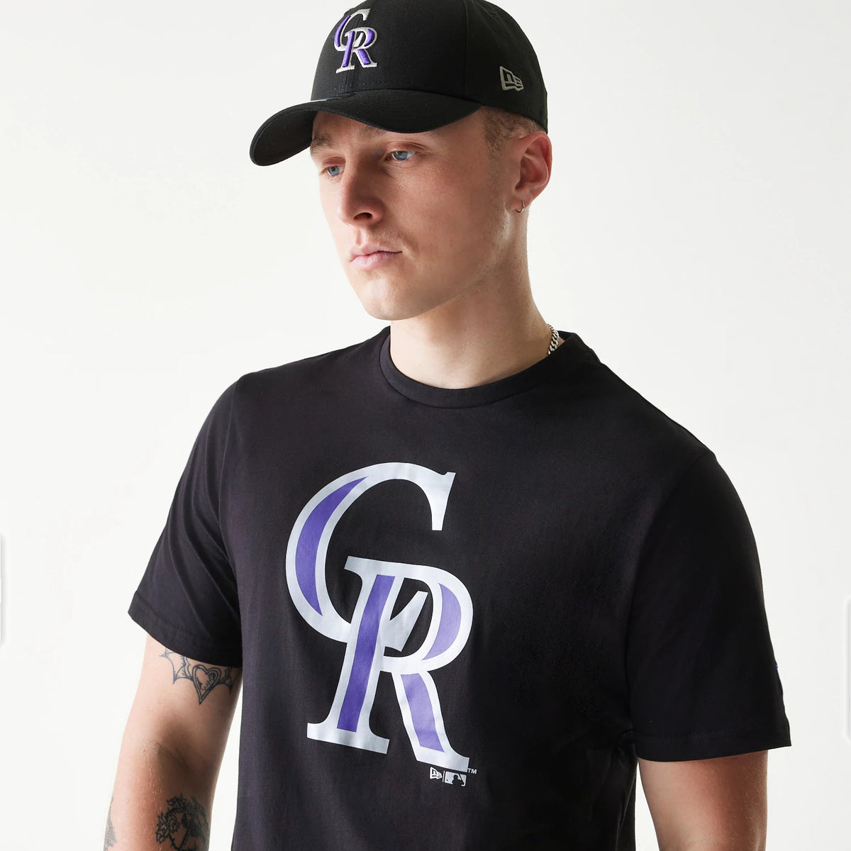 New Era Colorado Rockies MLB Regular Black T-Shirt, on-model view showing the large purple and white CR logo on the chest, worn alongside a black cap featuring a raised CR logo on the front and a debossed New Era logo on the side.