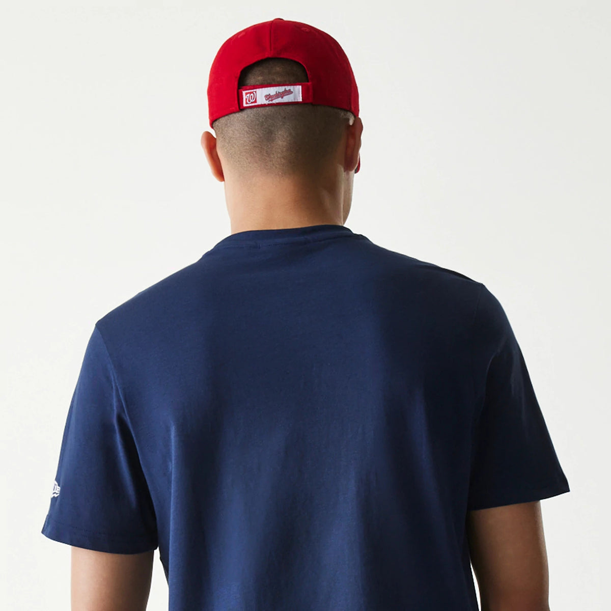 New Era Washington Nationals MLB Regular Dark Blue T-Shirt, rear view showing the plain dark blue back of the T-shirt worn by a person, the New Era logo embroidered on the left sleeve and a red cap with a white strap.