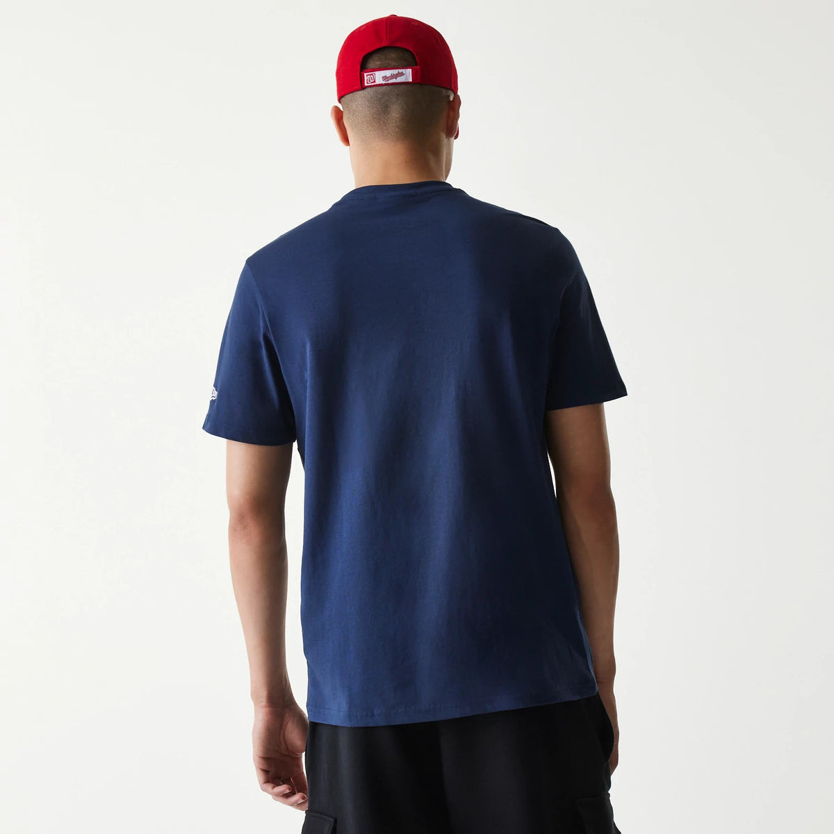 New Era Washington Nationals MLB Regular Dark Blue T-Shirt, rear view showing the plain dark blue fabric of the T-shirt, a debossed New Era logo on the left sleeve, worn by a person in a red cap facing away.