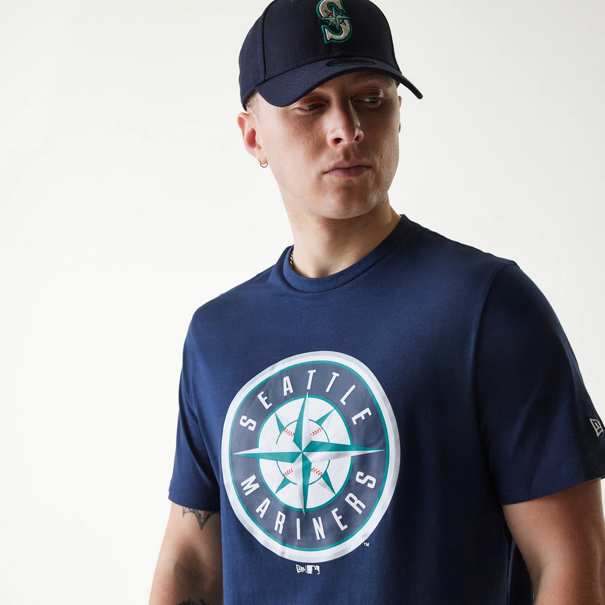 New Era Seattle Mariners MLB Regular Dark Blue T-Shirt, on-model view showing a large Seattle Mariners logo centred on the chest, worn by a person also wearing a dark blue cap with a raised team logo on the front.