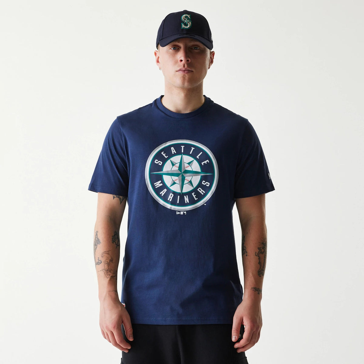 New Era Seattle Mariners MLB Regular Dark Blue T-Shirt, on-model view showing a large circular Seattle Mariners logo on the centre of the chest, a navy cap with a raised team emblem, and a small embroidered logo on the left sleeve.