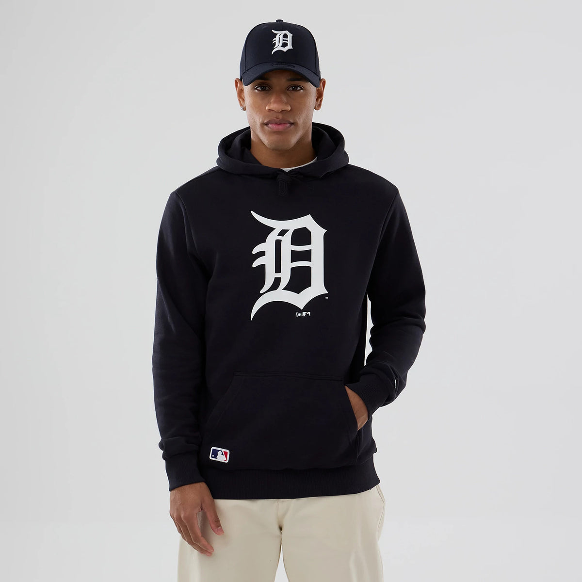 New Era Detroit Tigers MLB Navy Pullover Hoodie, on-model view showing a large white 'D' logo on the front centre, a classic pouch pocket, and a black cap with the same white 'D' logo worn by the model.