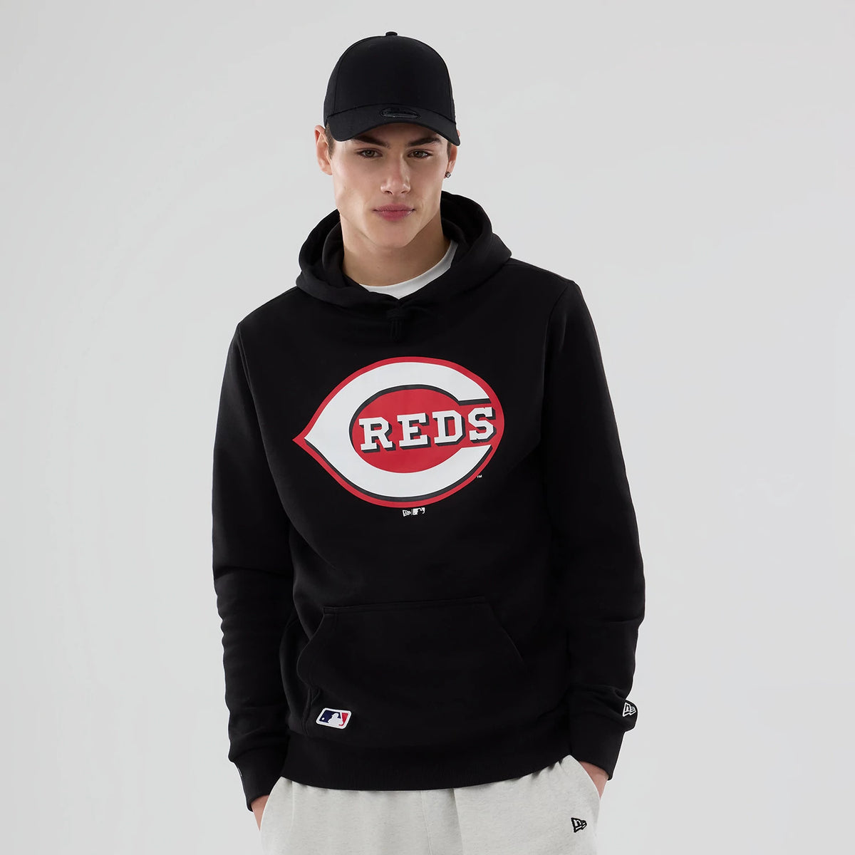 New Era Cincinnati Reds MLB Black Pullover Hoodie, on-model view showing a large red and white logo on the front centre, a kangaroo pocket below, New Era flag on the left sleeve, and a black cap with a debossed logo.
