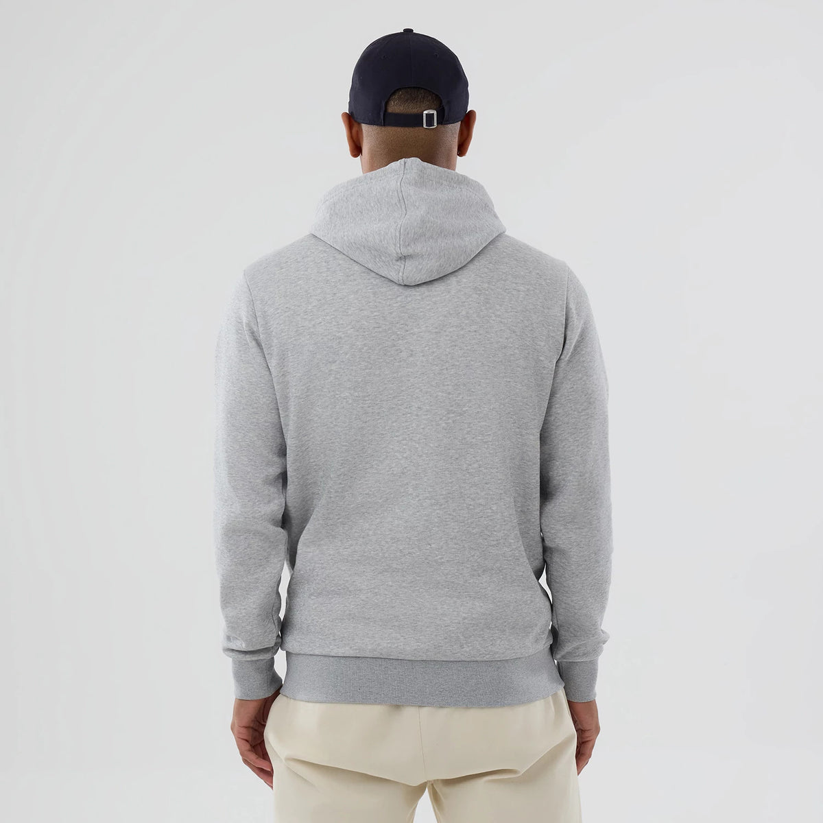 New Era Chicago Cubs MLB Grey Pullover Hoodie, rear view showing the plain grey back with an attached hood, ribbed cuffs and hem, worn by a person in beige trousers and a dark cap with an eyelet and strap at the back.