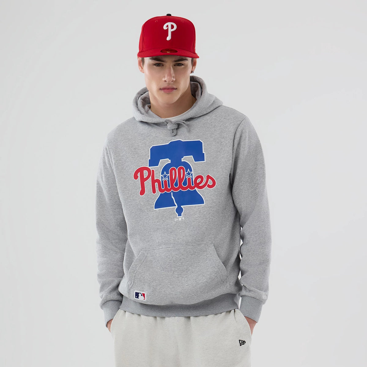 New Era Philadelphia Phillies MLB Grey Pullover Hoodie, on-model view showing the front graphic with a large blue bell and red Phillies text, a kangaroo pocket at the front, and a white tag near the hem above the grey ribbed cuffs.