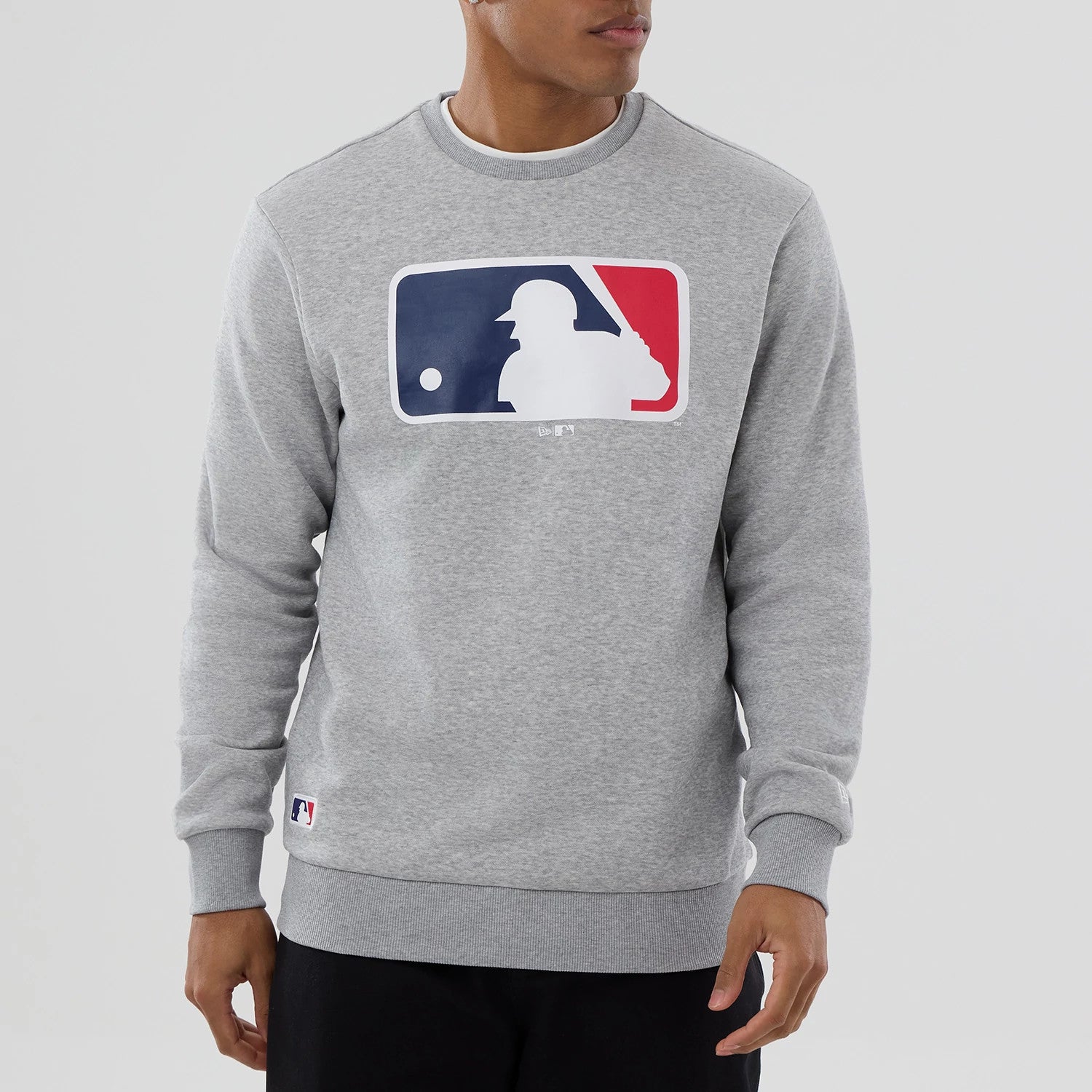 New Era MLB Generic Logo Grey Crew Neck Sweater, front view on a model featuring a large MLB logo in red, white, and blue centred on the chest, with ribbed cuffs and hem and a small MLB logo patch near the waistband.