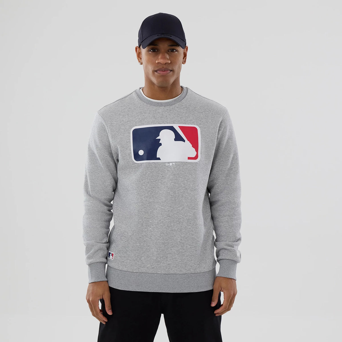 New Era MLB Generic Logo Grey Crew Neck Sweater, on a model wearing a navy cap, with a prominent MLB logo in red, white, and blue centred on the chest, and a small MLB logo label near the ribbed hem.