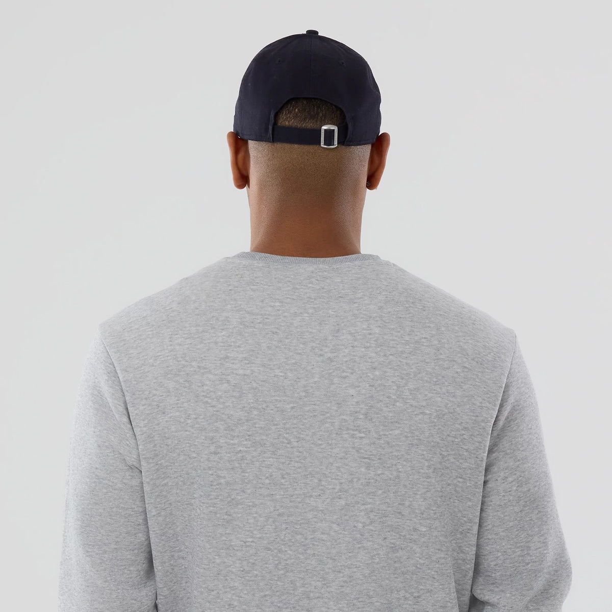 New Era MLB Generic Logo Grey Crew Neck Sweater, on-model rear view showing the plain design of the soft grey fabric, paired with a black cap that features an adjustable strap and metal buckle at the back.