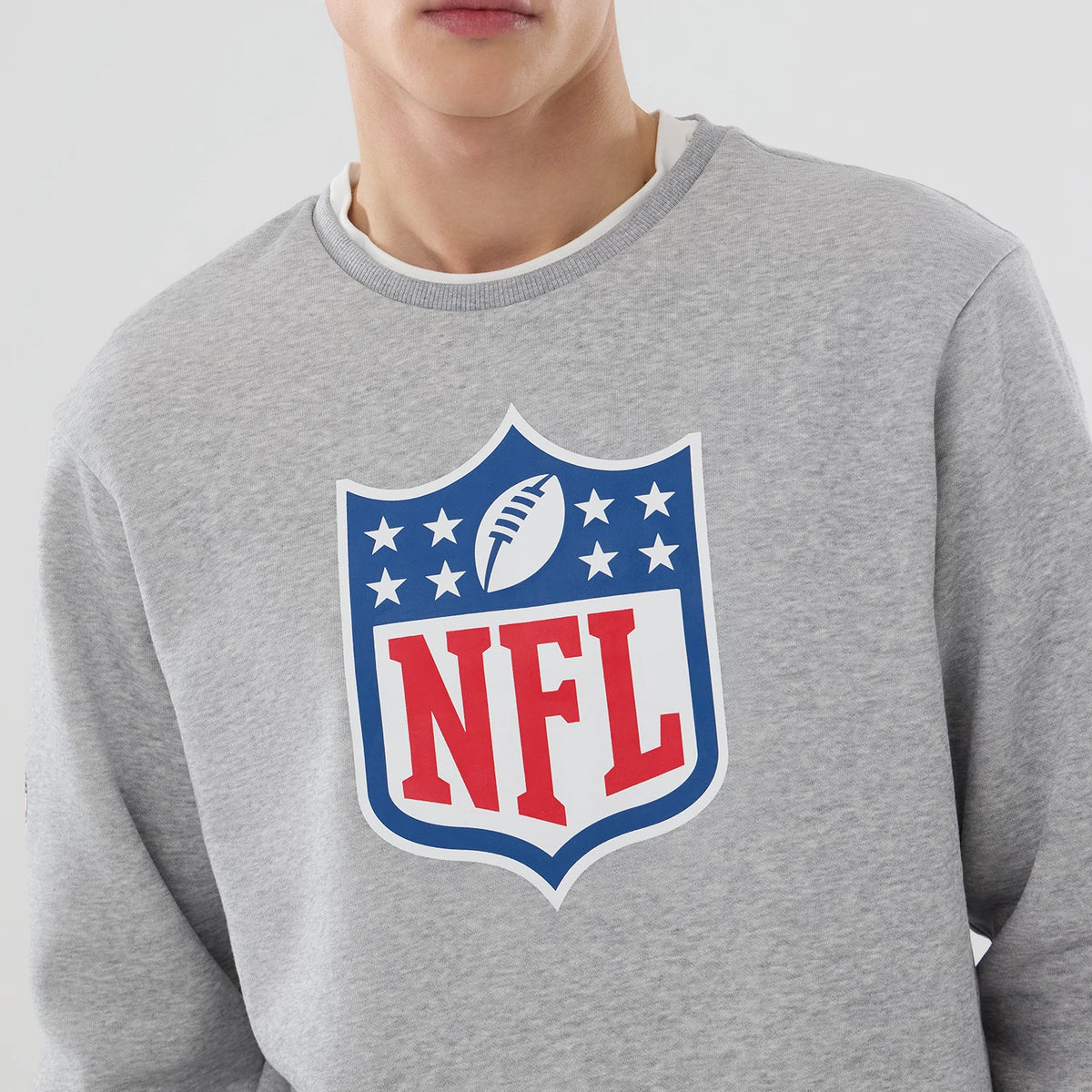 New Era NFL Generic Logo Grey Crew Neck Sweater, front view on a model showing the large printed red and blue NFL shield logo centred on the chest, with a ribbed collar and a glimpse of a white layer underneath.