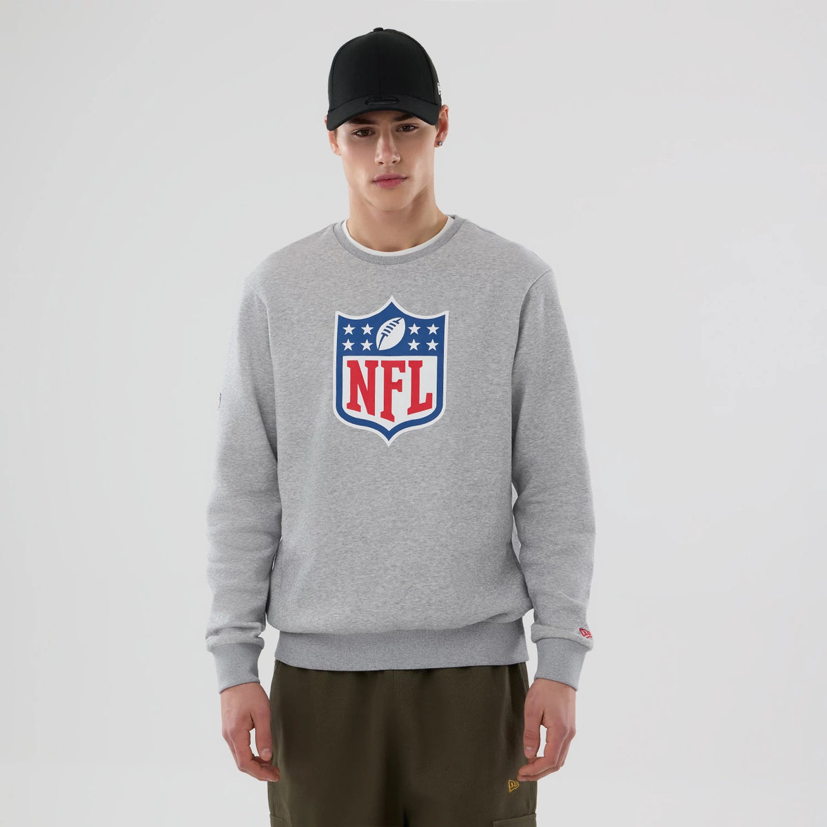 New Era NFL Generic Logo Grey Crew Neck Sweater, on-model front view with large NFL shield logo on the chest in red, white and blue, paired with olive trousers and a plain black cap.