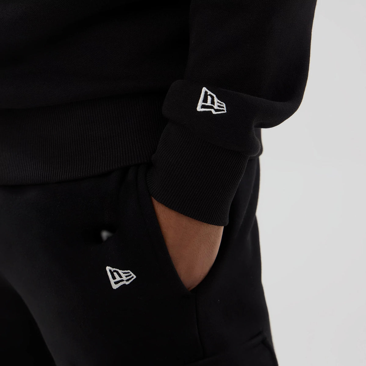 New Era NFL Generic Logo Black Crew Neck Sweater, close-up showing the white embroidered New Era flag logo on the ribbed cuff above the wrist and another logo near the pocket on matching black sweatpants.