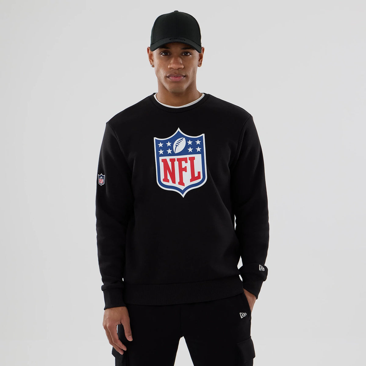 New Era NFL Generic Logo Black Crew Neck Sweater, front view on a model wearing a black cap, showing the large central NFL shield logo in red, blue and white on the chest and a smaller NFL logo on the left sleeve.