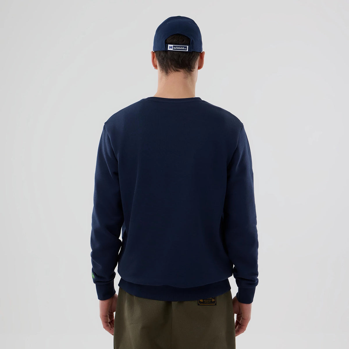 New Era Seattle Seahawks NFL Dark Blue Crew Neck Sweater, back view on a model wearing olive trousers, showing the plain dark blue fabric with ribbed cuffs and hem, alongside a matching dark blue cap.