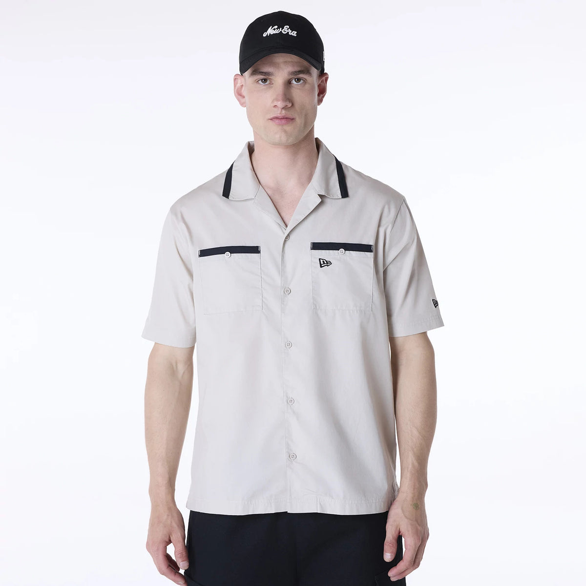 New Era Revere Cream Short Sleeve Shirt, front view on a model wearing a black cap, featuring black piping on the collar and pockets, button closure, and a small embroidered flag logo on the left chest.