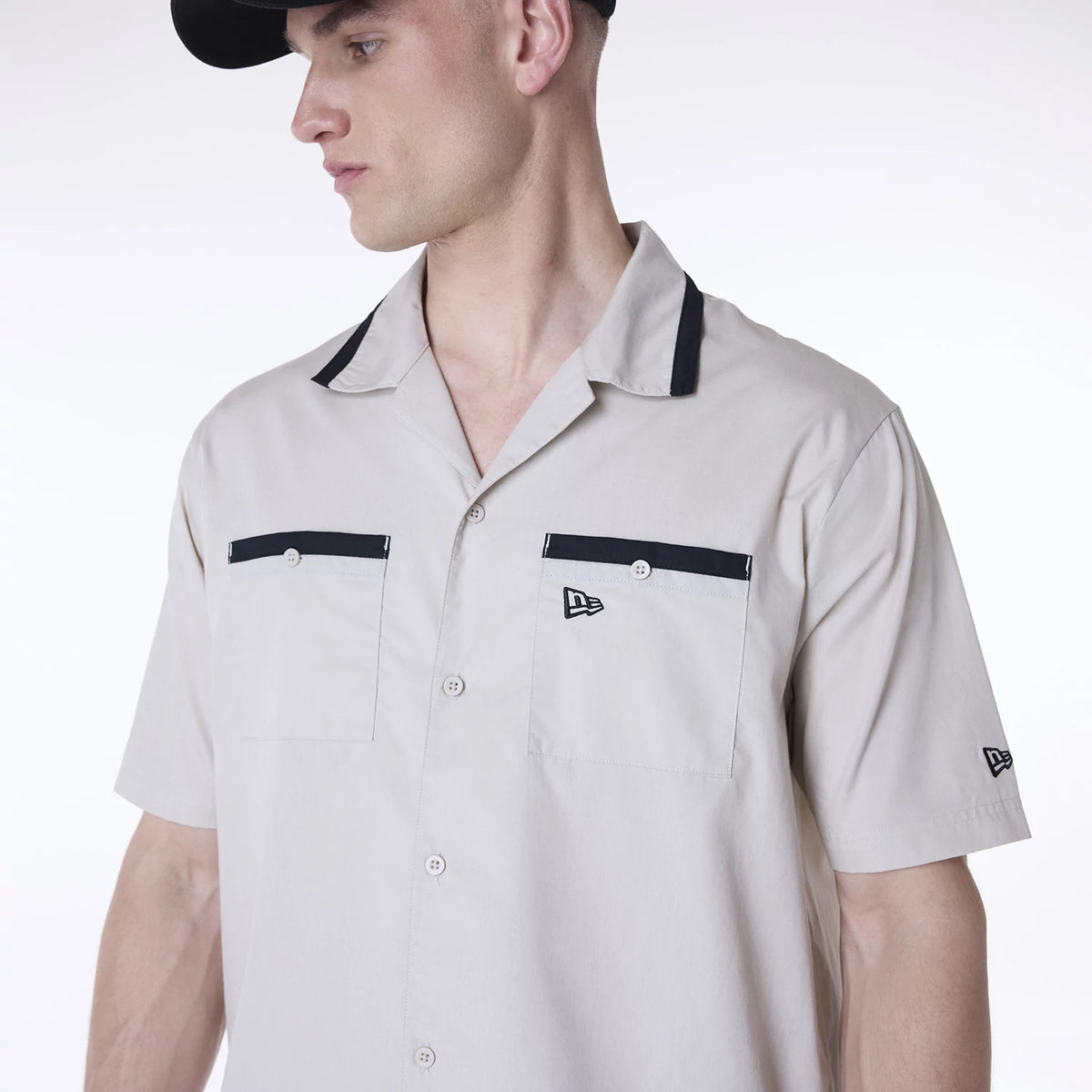 New Era Revere Cream Short Sleeve Shirt, on a model showing black trim on the collar and pocket flaps with a New Era flag logo on the chest and sleeve, styled with a black cap, highlighting casual summer attire.