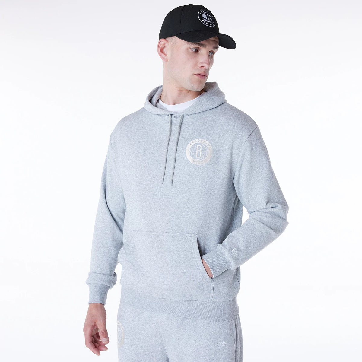 New Era Brooklyn Nets NBA League Essential Grey Oversized Pullover Hoodie, on-model view showing the grey hoodie with a raised Brooklyn Nets logo on the left chest, a front pocket, and a drawstring hood, paired with matching grey sweatpants.