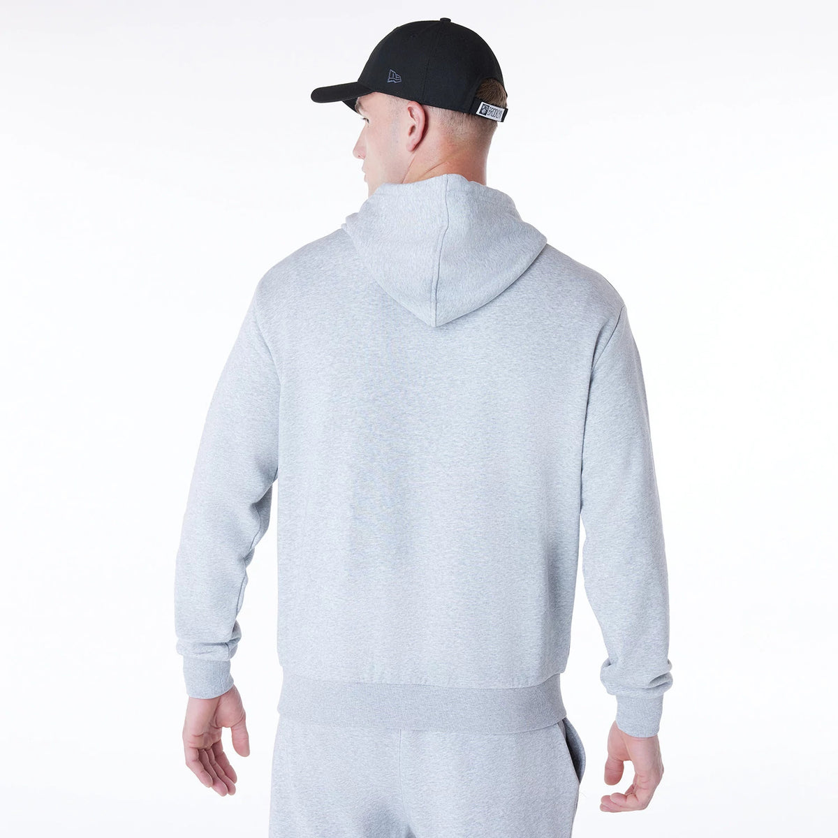 New Era Brooklyn Nets NBA League Essential Grey Oversized Pullover Hoodie, rear view showing the plain grey fabric with a generously sized hood, ribbed cuffs and hem, worn by a model in matching grey trousers also wearing a black cap.