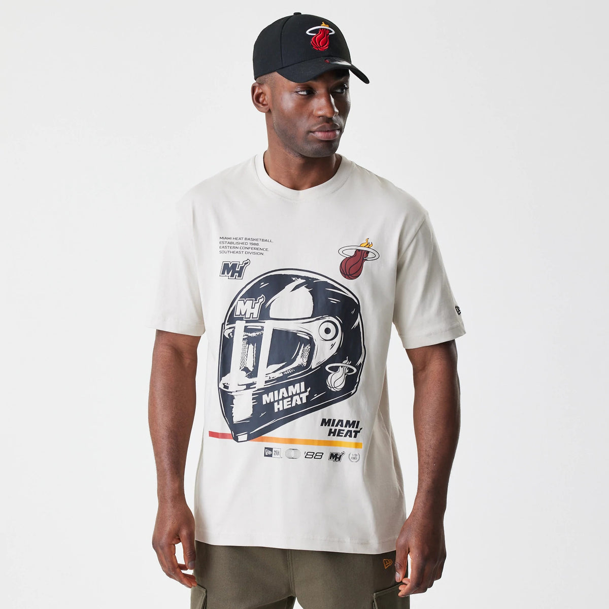 New Era Miami Heat Modern Driver Light Beige T-Shirt, on-model view showing a graphic of a helmet on the front, small Miami Heat logo on the chest, and New Era logo on the left sleeve, light beige fabric with short sleeves.