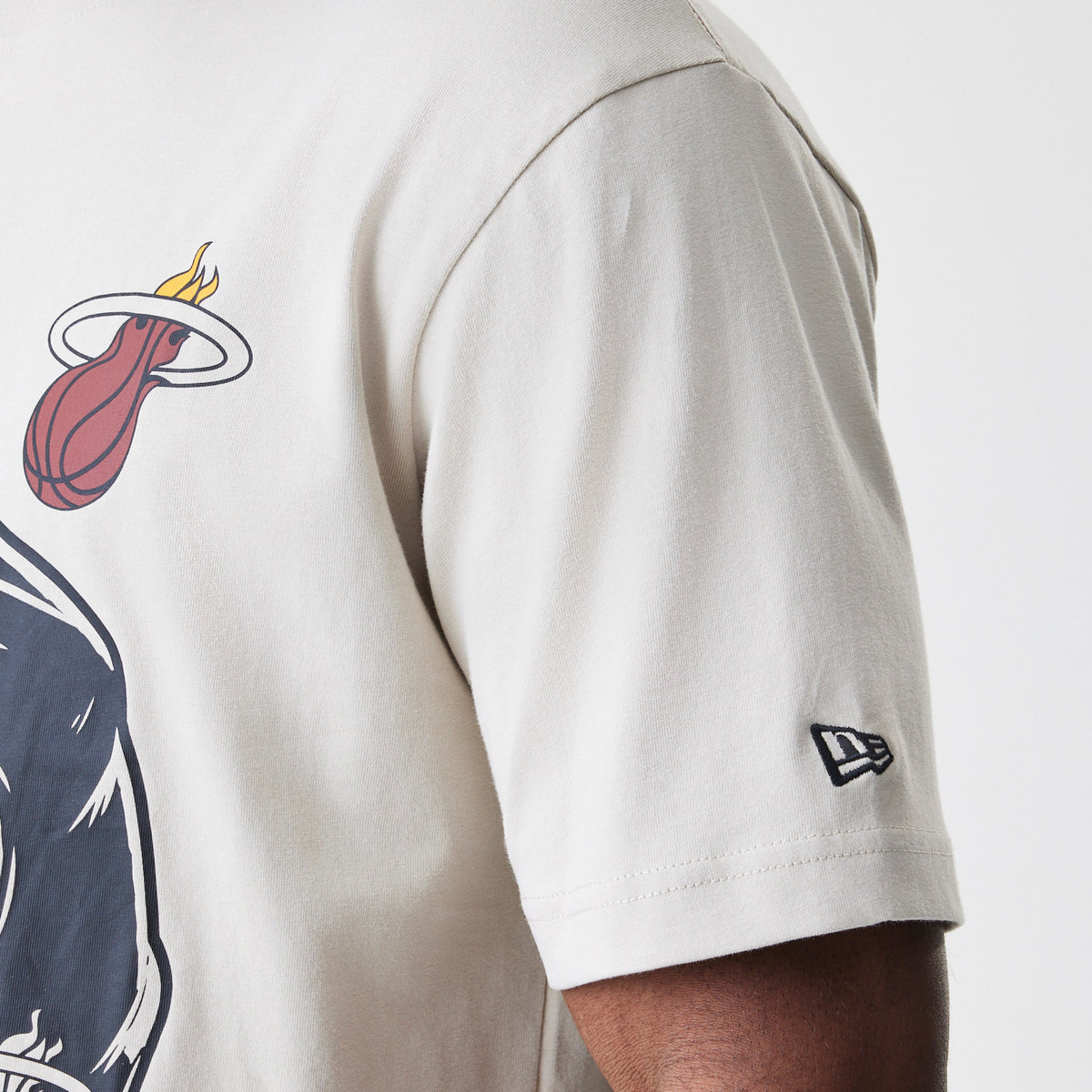 New Era Miami Heat Modern Driver Light Beige T-Shirt, side view showing a light beige short sleeve with a raised New Era logo near the cuff and a graphic of the Miami Heat flaming basketball emblem on the upper back.
