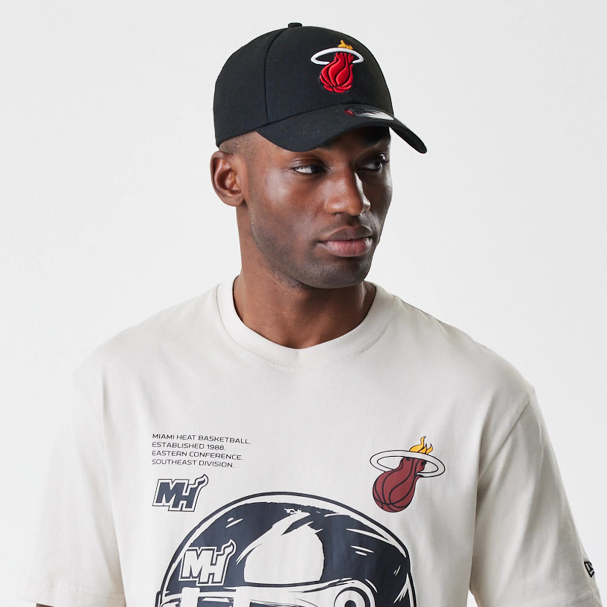 New Era Miami Heat Modern Driver Light Beige T-Shirt, on-model view showing the light beige fabric with a large black and white graphic and red Miami Heat logo on the chest, paired with a black cap featuring an embroidered Miami Heat logo.