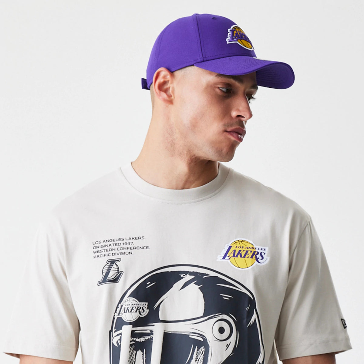 New Era LA Lakers Modern Driver Light Beige T-Shirt, on-model view with a large graphic of a driver's helmet in black on the front, a purple debossed Lakers logo on the chest and small text above the graphic.