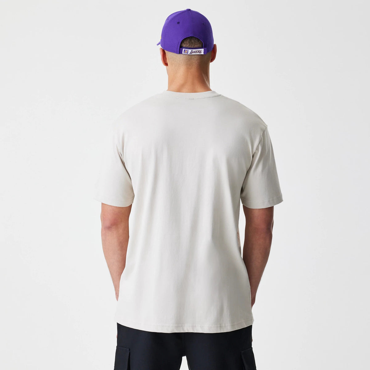 New Era LA Lakers Modern Driver Light Beige T-Shirt, on-model rear view showing the plain light beige back of the t-shirt with short sleeves, paired with a purple cap featuring a debossed LA Lakers logo on the back strap.